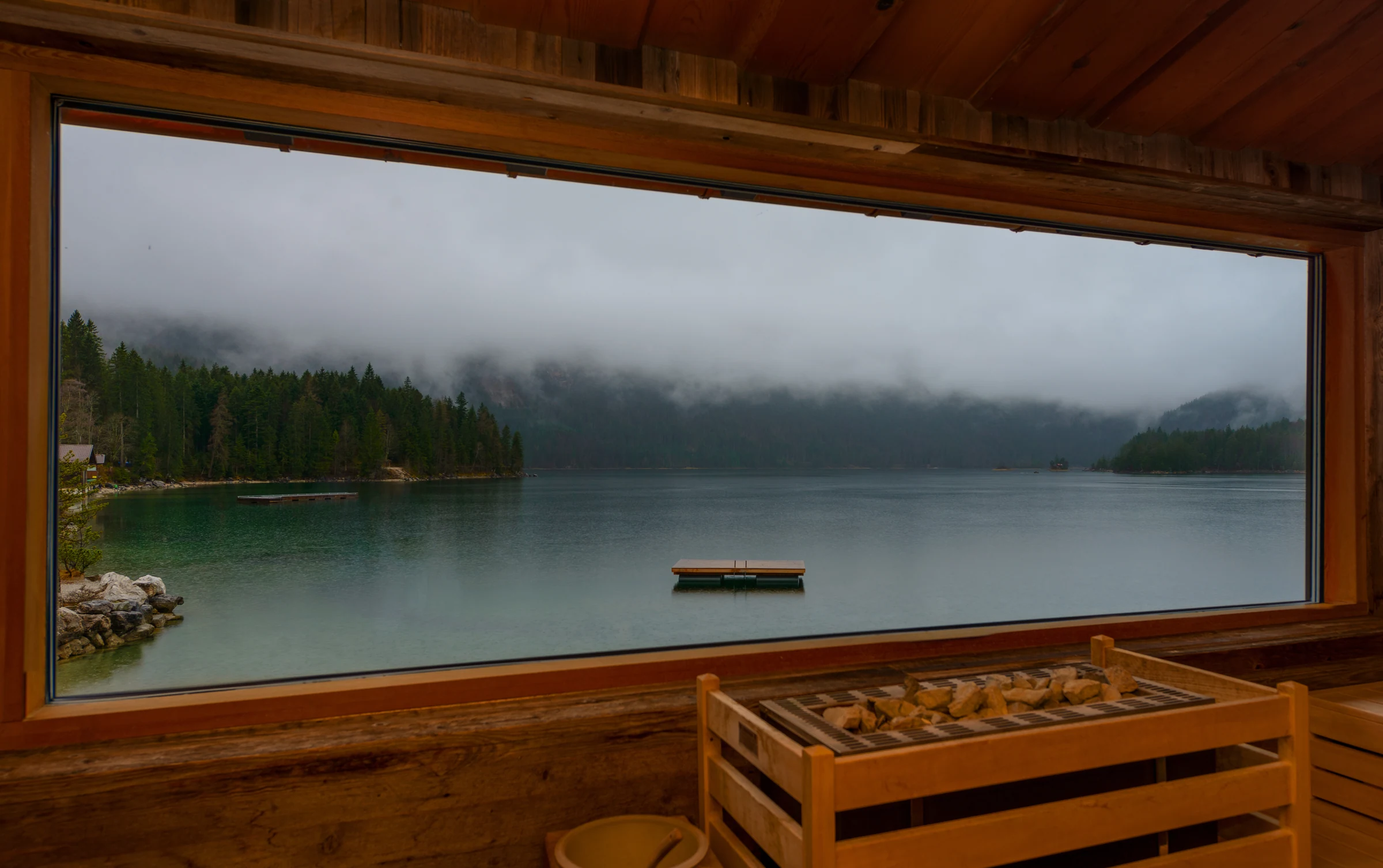 A timber window frames the lake and wooded slopes of Eibsee beneath low cloud.